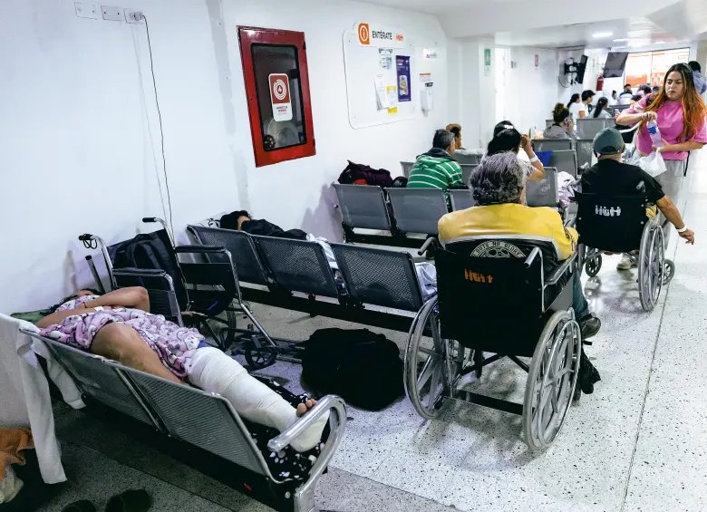 Personas esperan en sillas y sillas de ruedas dentro de una sala de urgencias. Referencia a deudas de la EPS.