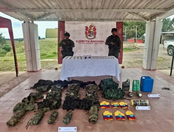Dos uniformados del Ejército detrás de una mesa, con prendas camufladas, equipos y recipientes exhibidos en el suelo.